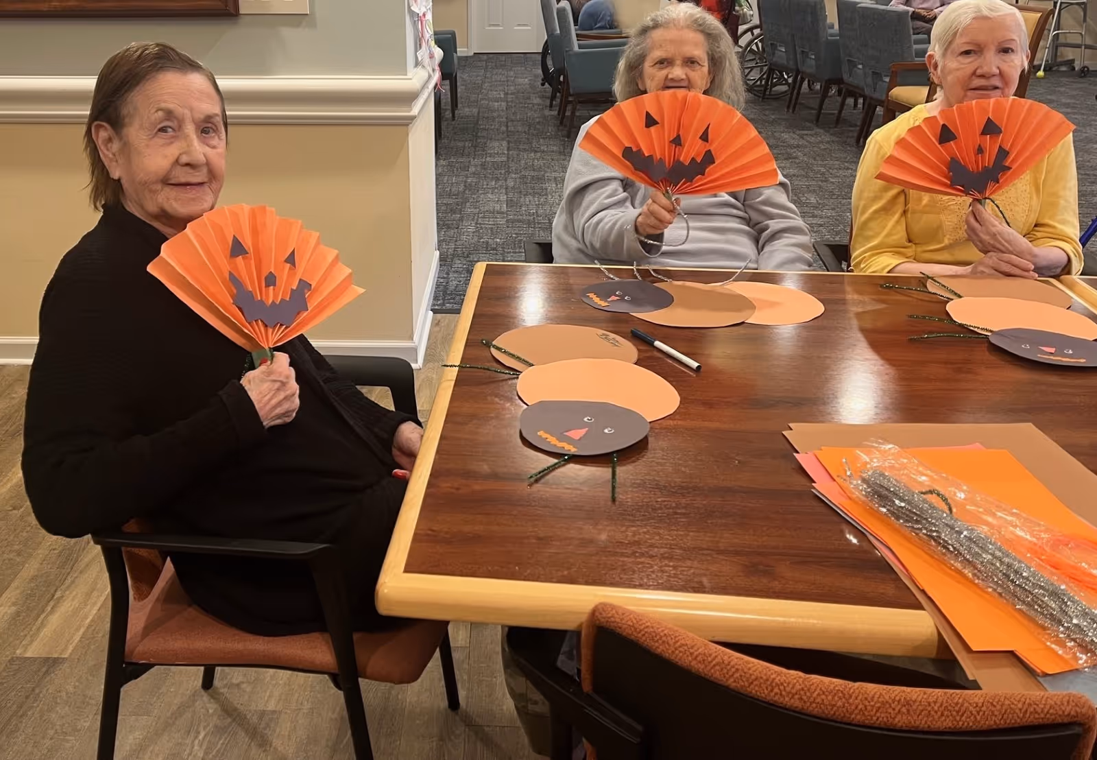 Three elderly women sitting around a wooden table in a common area, each holding an orange paper fan decorated with a jack-o'-lantern face. On the table are craft materials including paper cutouts and pipe cleaners shaped like pumpkins.
