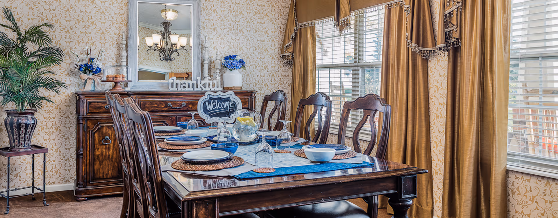 A warmly decorated dining room with a wooden dining table set for six people, featuring blue and white dishware, woven placemats, and a pitcher of water with lemon. Behind the table is a wooden sideboard with a large mirror, a vase with blue flowers, and a decorative sign that says 'thankful'. The room has patterned wallpaper, large windows with golden curtains, and a potted plant on a small table.
