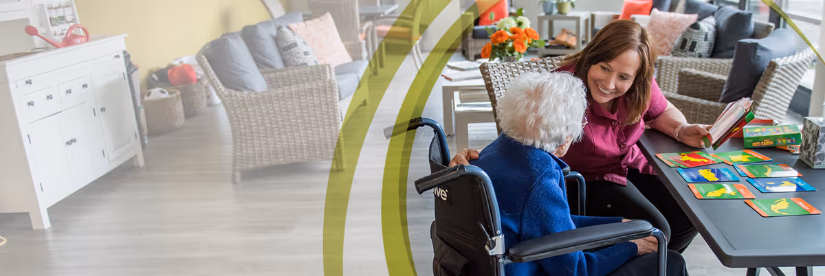 Caregiver smiling and playing a card game with an elderly resident in a wheelchair in a bright communal living room with wicker seating and a sideboard.
