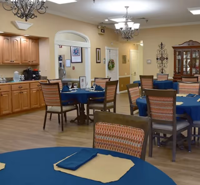 Dining room with round tables set with blue tablecloths and wooden chairs, chandeliers, and a serving cabinet.