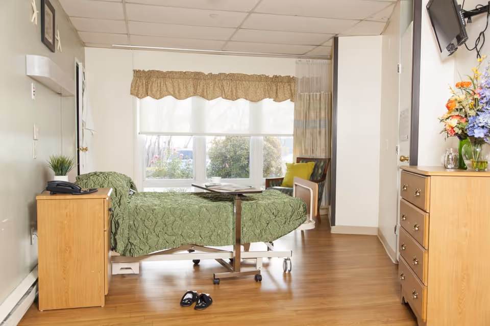 A well-lit room with a hospital-style bed covered in a green quilt, a wooden nightstand with a phone and plant, a small overbed table with a cup and papers, a wooden dresser with a vase of colorful flowers, a chair with a green cushion, and large windows with beige curtains letting in natural light.