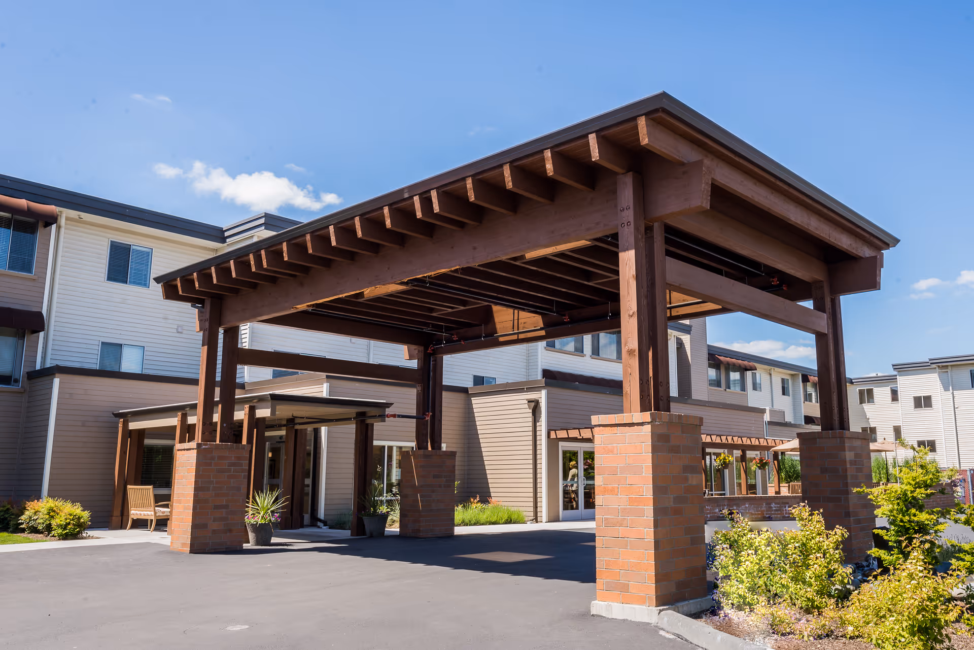 Entrance area of Village Concepts of Auburn - Brannan Park featuring a large wooden pergola supported by brick pillars, with the multi-story building in the background under a clear blue sky.
