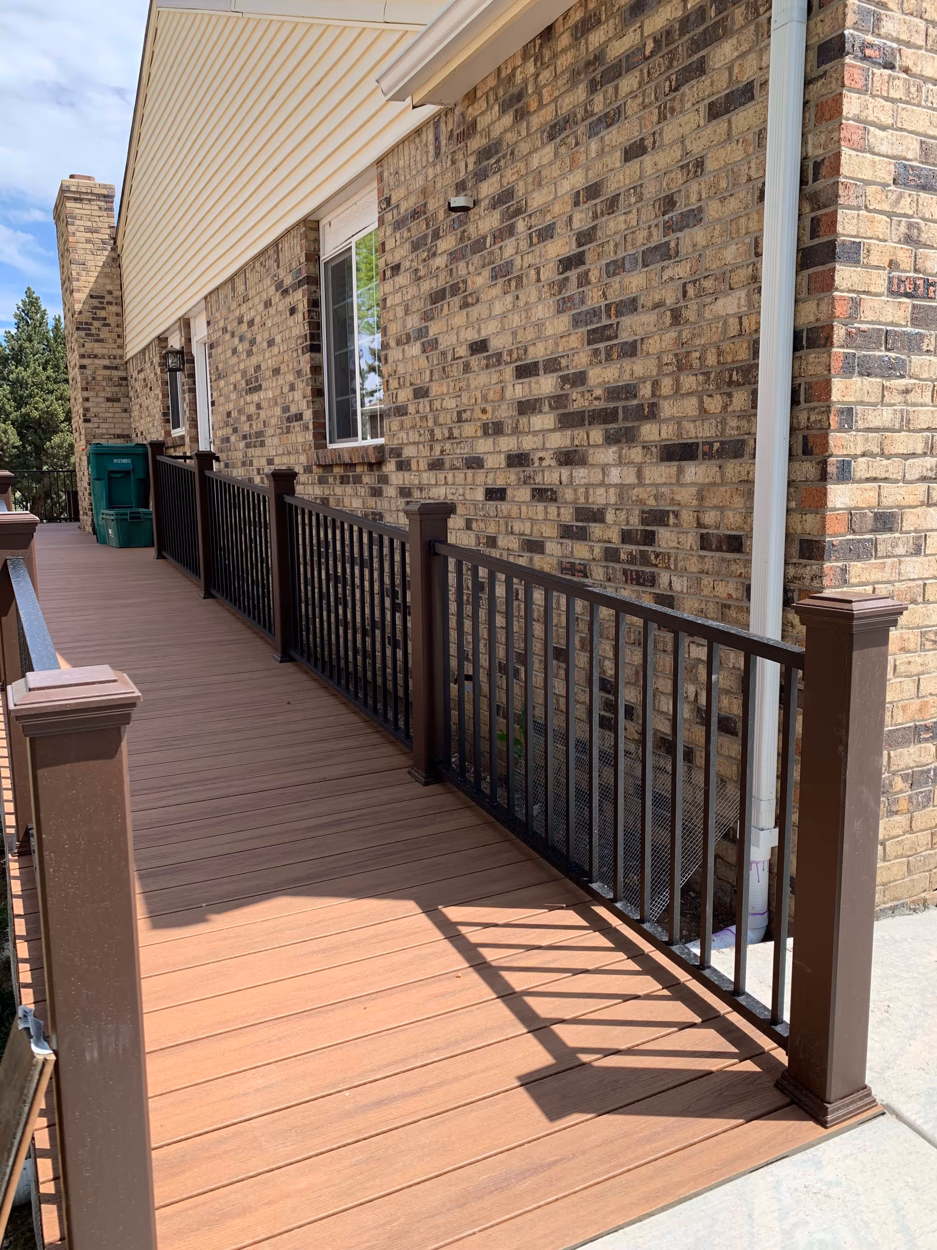 A wooden deck with brown railings attached to a brick building exterior. There is a window on the brick wall and a green trash bin at the far end of the deck. The sky is partly cloudy and some trees are visible in the background.