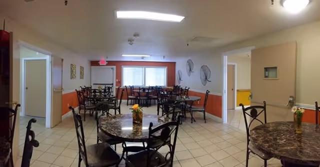 Dining area with round marble-top tables and metal chairs on a tiled floor, windows at the far wall and decorative wall fans.