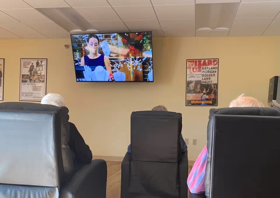 Three elderly individuals sitting in black recliner chairs facing a wall-mounted television screen in a common room. The TV displays a woman in a dark sleeveless top with holiday decorations in the background. The beige walls have framed vintage posters, including one for The Wizard of Oz.