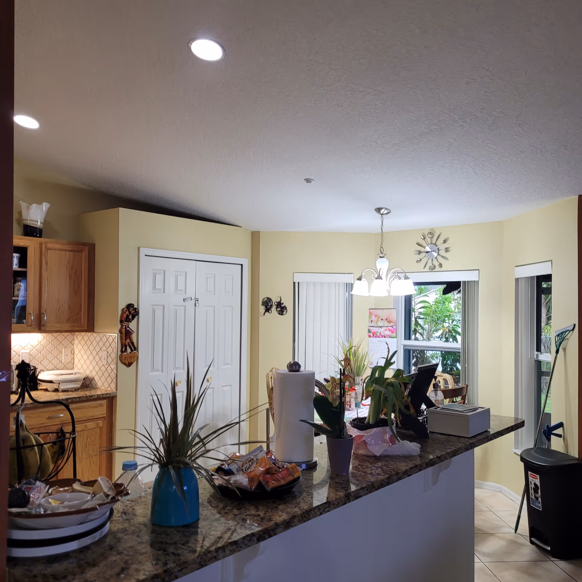 Open kitchen and dining area with a granite countertop island topped with potted plants and snacks, hanging light fixture, cabinets, and windows with vertical blinds.