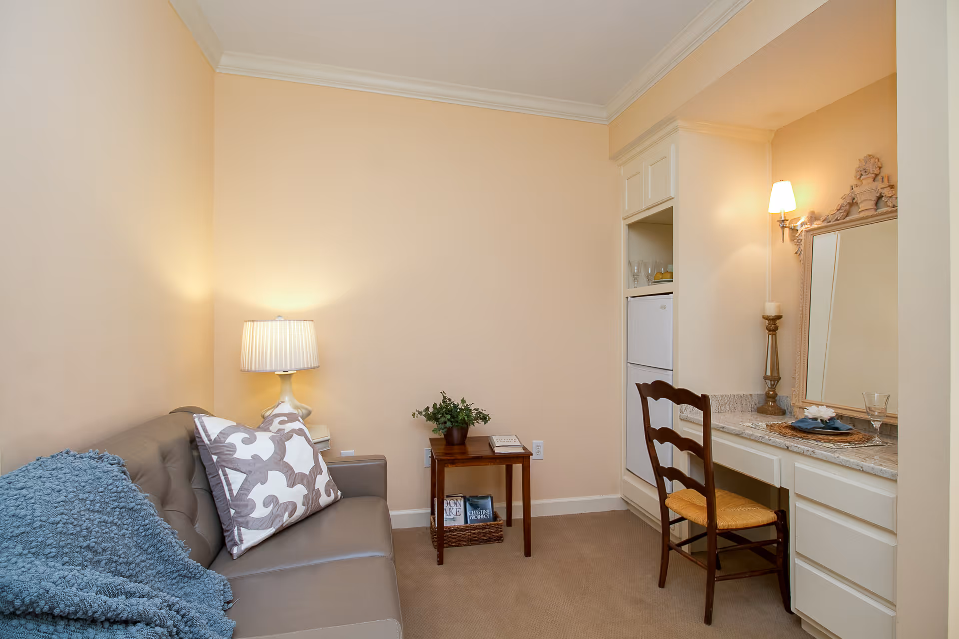 A cozy interior room with a beige leather sofa adorned with a patterned pillow and a blue throw blanket. Next to the sofa is a small wooden side table with a potted plant and books underneath. Across from the sofa is a built-in vanity desk with a wooden chair, a decorative mirror, a candle holder, and a wall-mounted lamp providing warm lighting. The walls are painted light beige and the floor is carpeted.