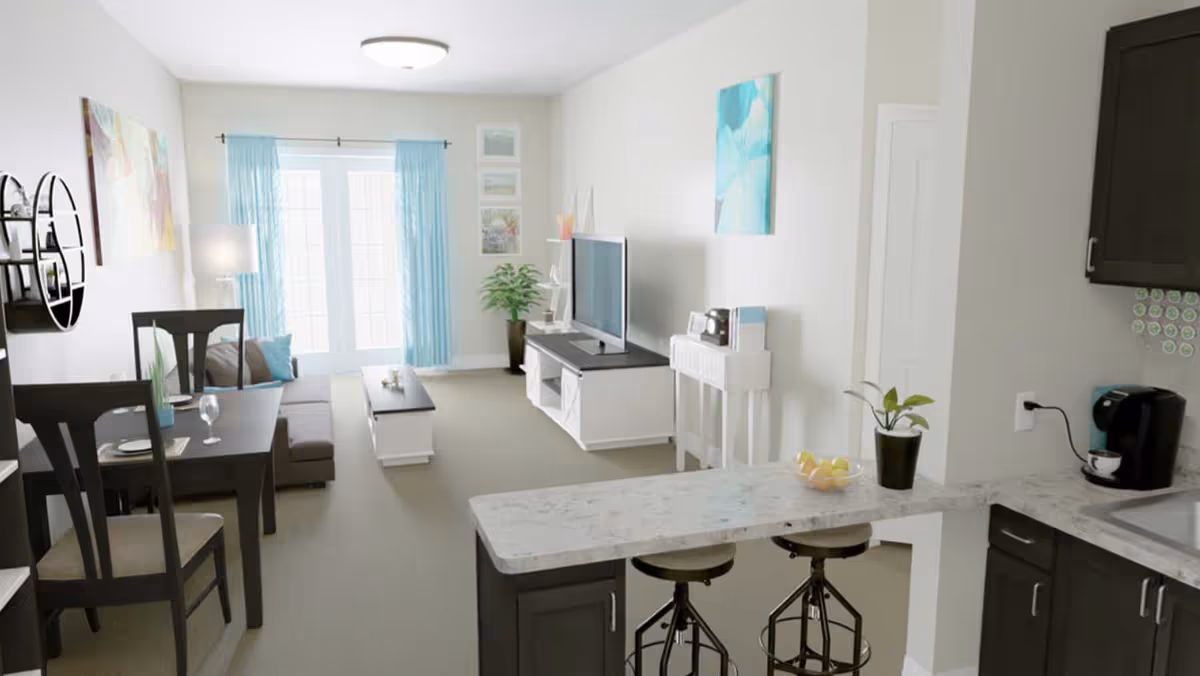 A bright and modern living area in a senior living facility featuring a dining table with chairs, a sofa with cushions, a coffee table, a TV on a white stand, and a kitchen counter with two stools. The room has light-colored walls, blue curtains on large windows, and decorative artwork on the walls.