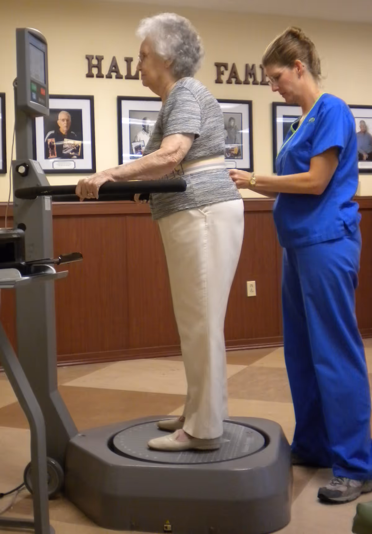 An elderly woman stands on a vibration exercise platform holding onto the handles, while a healthcare worker in blue scrubs assists her by adjusting a belt around her waist. The setting appears to be an indoor rehabilitation or therapy room with framed photos on the wall behind them.