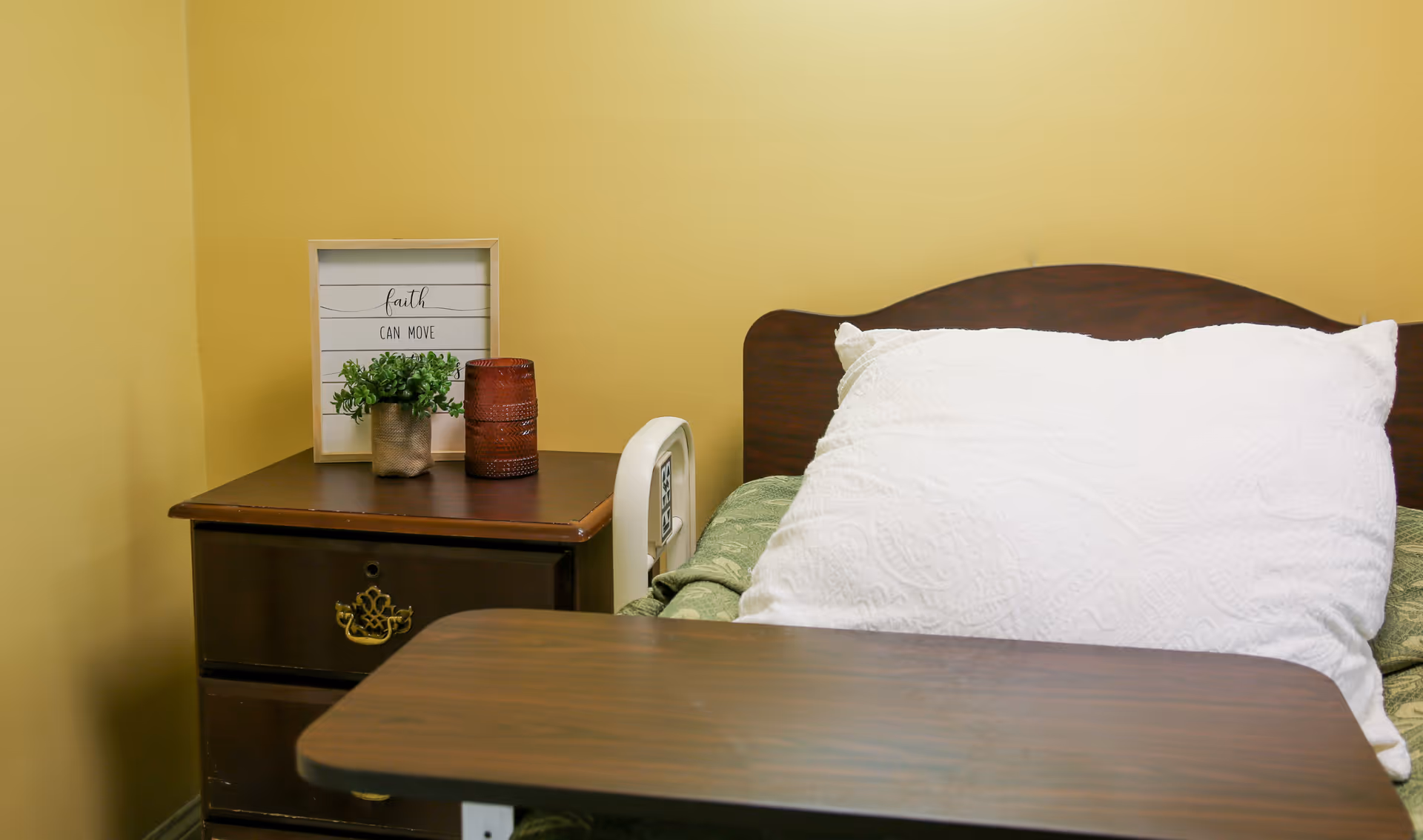Hospital-style bed with a large white pillow and wooden headboard next to a nightstand holding a potted plant and decorative sign in a softly lit room.