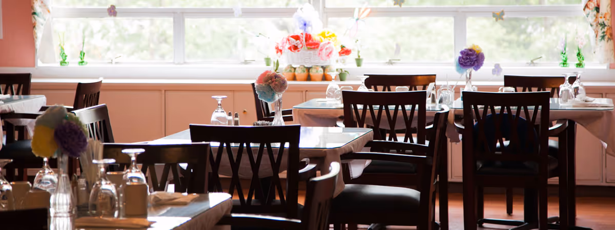 Bright dining room with multiple set tables and wooden chairs arranged by a large window decorated with flowers.