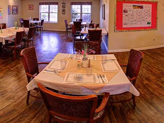 A dining room with several tables covered with beige tablecloths and set with placemats, napkins, utensils, and condiments. The room has wooden flooring, large windows with curtains, and a bulletin board on the wall.