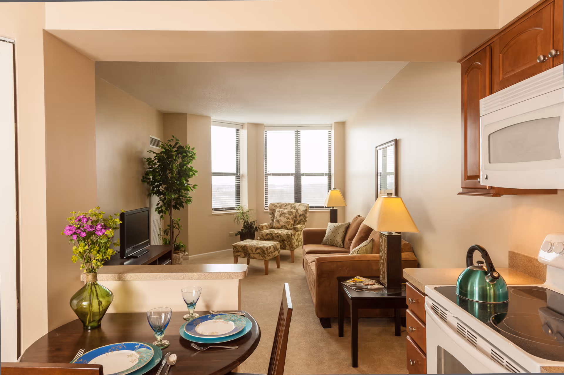 View of a cozy living area in a senior living facility with a small dining table set for two in the foreground, a kitchen area with a stove and microwave on the right, and a living room with a brown sofa, patterned armchair with ottoman, side table with lamp, and a TV on a stand near a window in the background.