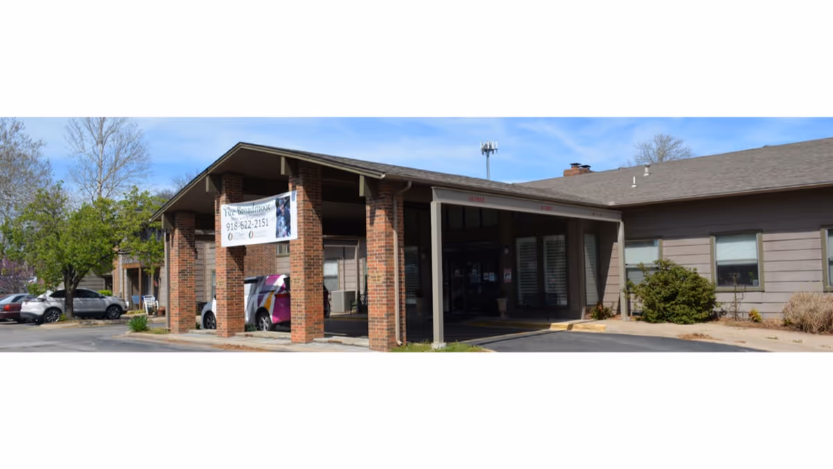 Exterior view of The Broadmoor Retirement Community building entrance with a covered drop-off area supported by brick columns. Several cars are parked nearby, and trees and shrubs surround the building under a clear blue sky.