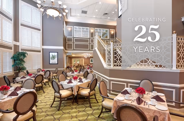 Elegant dining room with multiple round tables set with beige tablecloths, white plates, cups, silverware, and burgundy napkins. Each table has a centerpiece with red flowers. The room features high ceilings, large windows with white shutters, decorative wall paneling, and a chandelier. A staircase with ornate railing is visible in the background. Text overlay reads 'Celebrating 25 Years'.