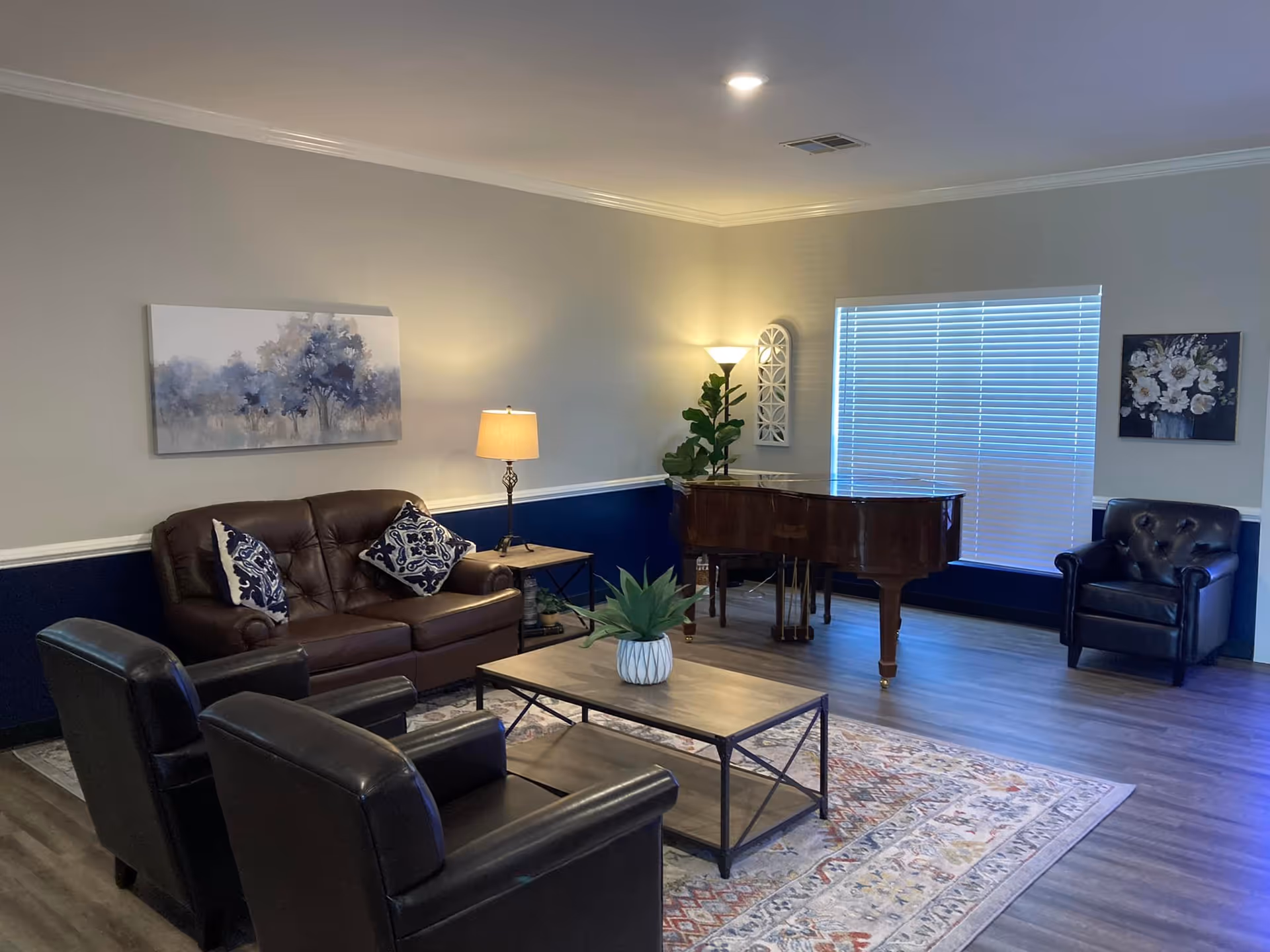 Well-lit living room with leather sofa and chairs arranged around a coffee table, a grand piano near a window, and wall art.
