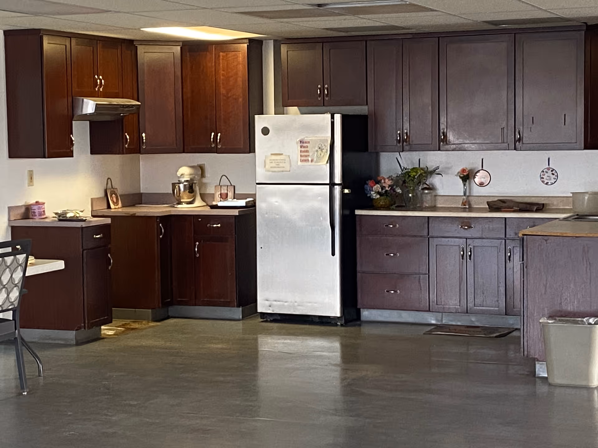Community kitchen with dark wood cabinets, a stainless steel refrigerator, countertops with a mixer and flowers, and a table at the left.
