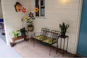 A small outdoor seating area with a black metal bench featuring colorful floral cushions, a small wooden table with potted plants, and a tall plant stand with a green plant. The wall behind is white with horizontal siding, decorated with colorful flower wall hangings and a small birdhouse. A blue door is visible on the right side.