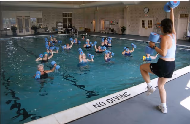 A group of elderly people participating in a water exercise class in an indoor swimming pool, following an instructor who is standing poolside holding blue foam dumbbells.