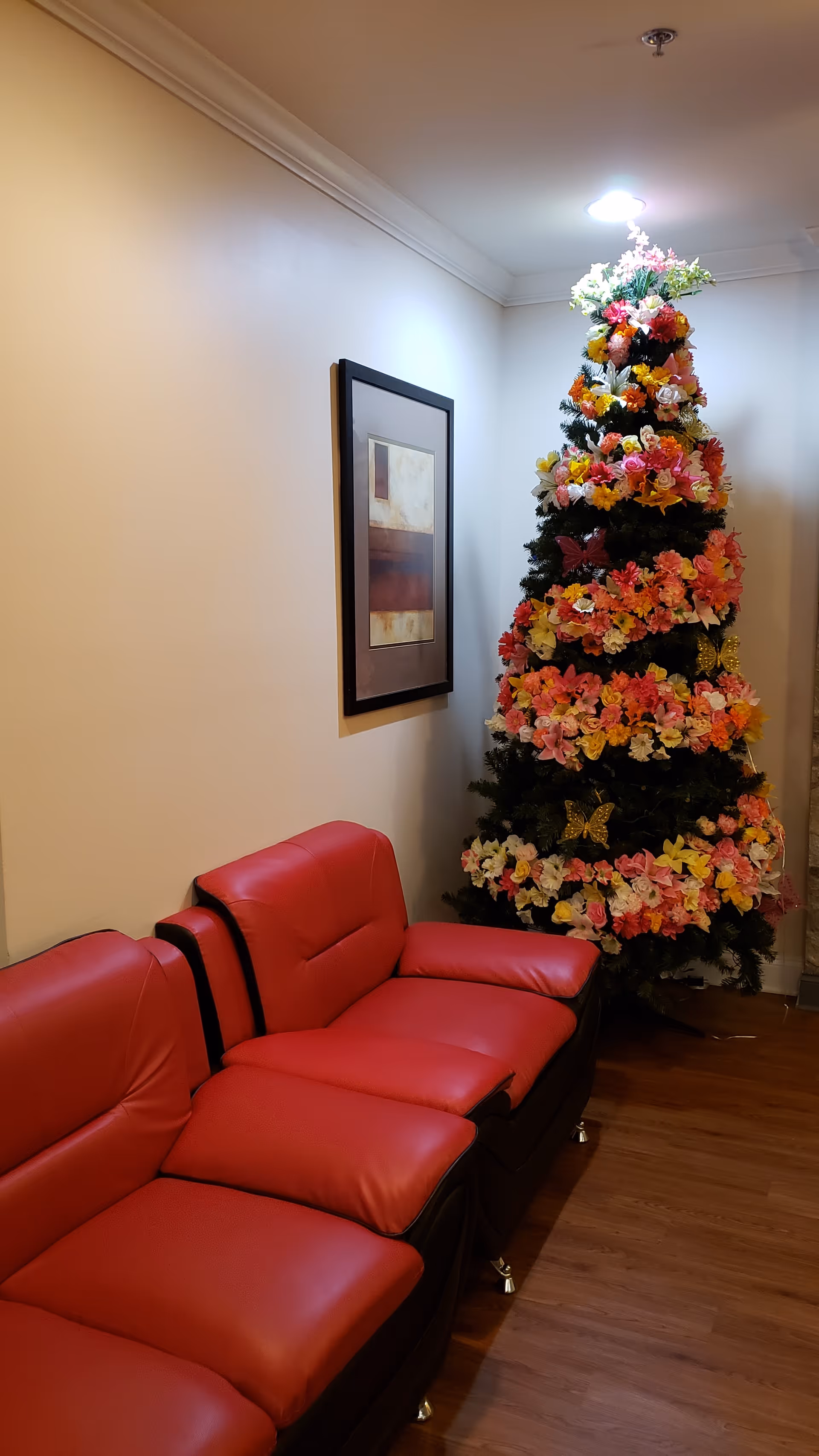 A cozy corner of a room featuring a red leather sofa with three seats and a tall Christmas tree decorated with numerous colorful flowers and butterfly ornaments. A framed abstract painting hangs on the wall above the sofa, and the floor is wooden.