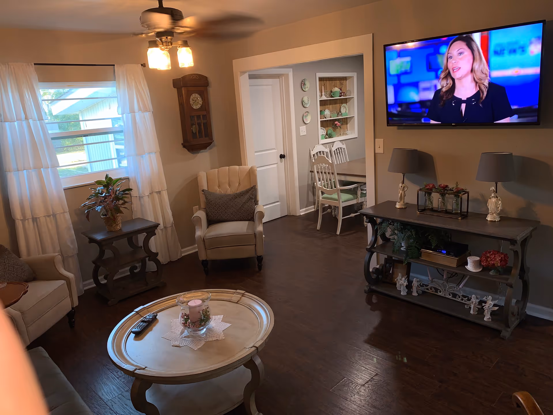 A cozy living room with wooden flooring, beige armchairs, a round coffee table with a candle centerpiece, a wall-mounted TV showing a woman speaking, a wooden console table with lamps and decorative items, a wall clock, and a window with white curtains letting in natural light. A dining area with white chairs and a table is visible through an open doorway.