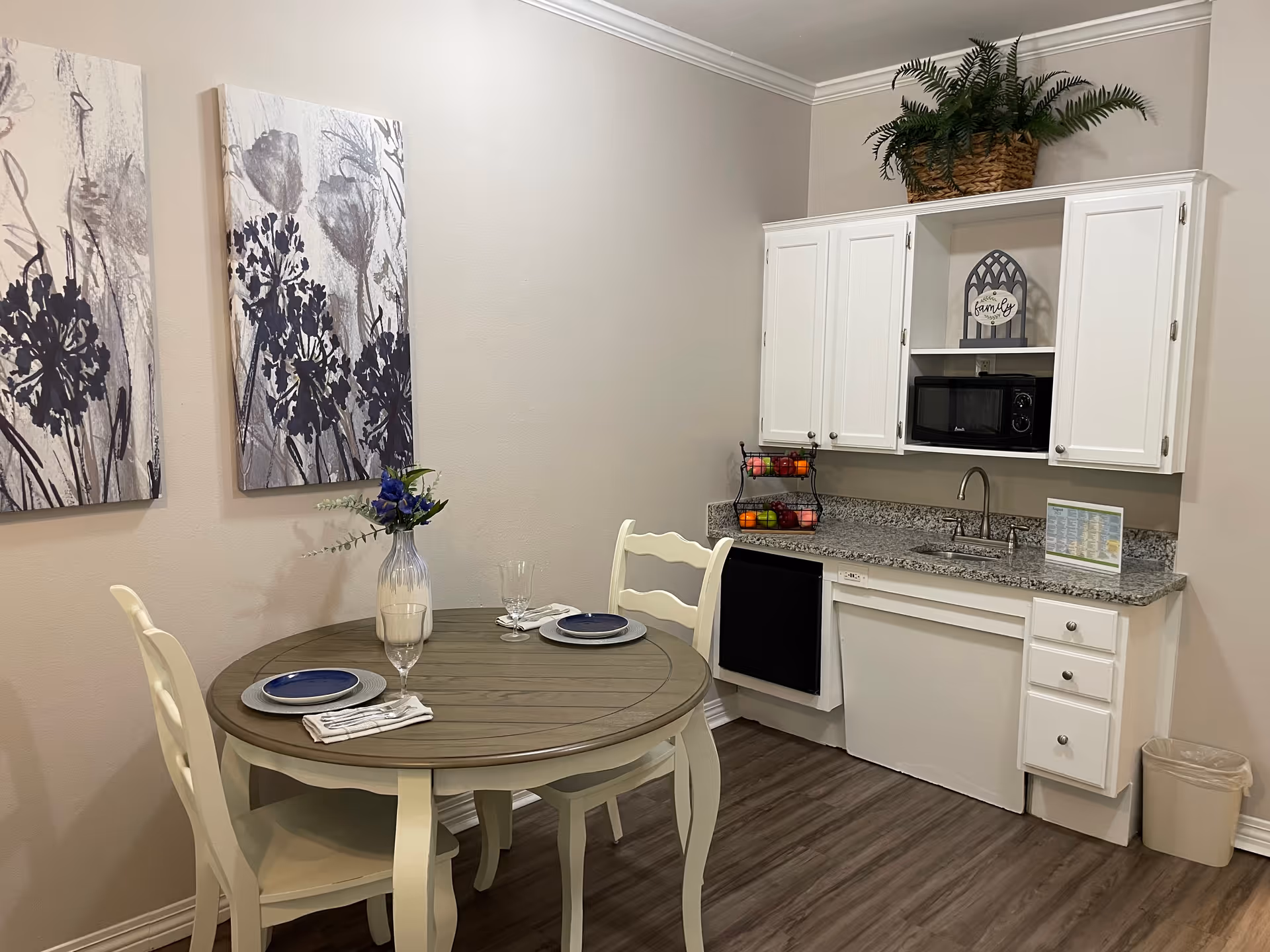 A small dining area with a round wooden table set for two with blue plates, glasses, and napkins. Behind the table is a kitchenette with white cabinets, a granite countertop, a microwave, a sink, and a two-tier fruit basket. Two floral paintings hang on the wall above the table, and a potted plant sits on top of the cabinets.