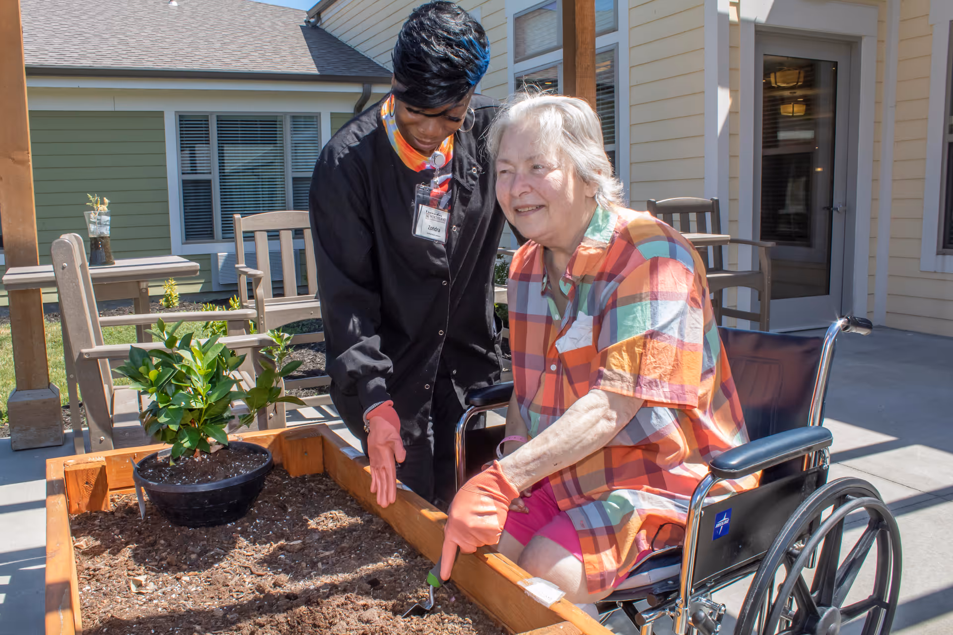 An elderly woman in a wheelchair wearing a colorful plaid shirt and pink shorts is gardening with the assistance of a caregiver dressed in black. They are outdoors near a raised garden bed with soil and a potted plant, surrounded by chairs and a building with windows and a door.