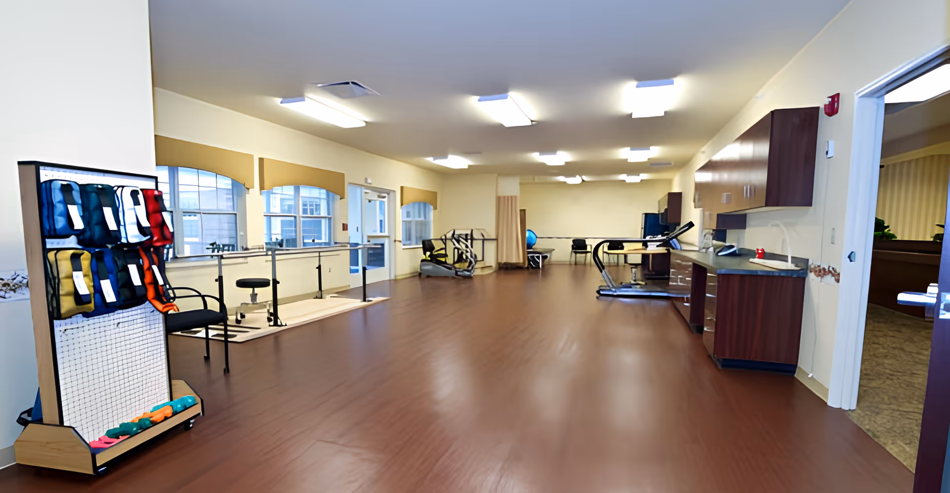 A spacious rehabilitation or physical therapy room with wooden flooring, exercise equipment including a treadmill, parallel bars for walking practice, weights, and a stationary bike. The room has large windows with beige valances, overhead fluorescent lighting, and a countertop with cabinets along one wall. An open door leads to an adjacent room.
