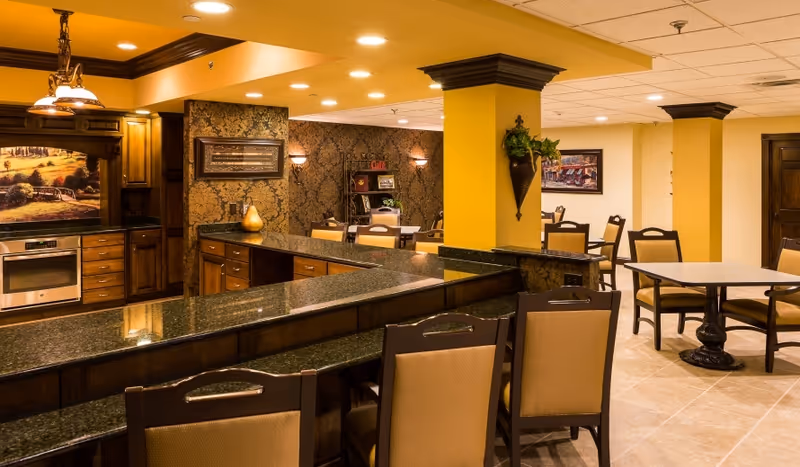 Interior view of a senior living facility dining area and kitchen with granite countertops, wooden cabinetry, multiple tables and chairs, warm yellow walls, and decorative wall art.