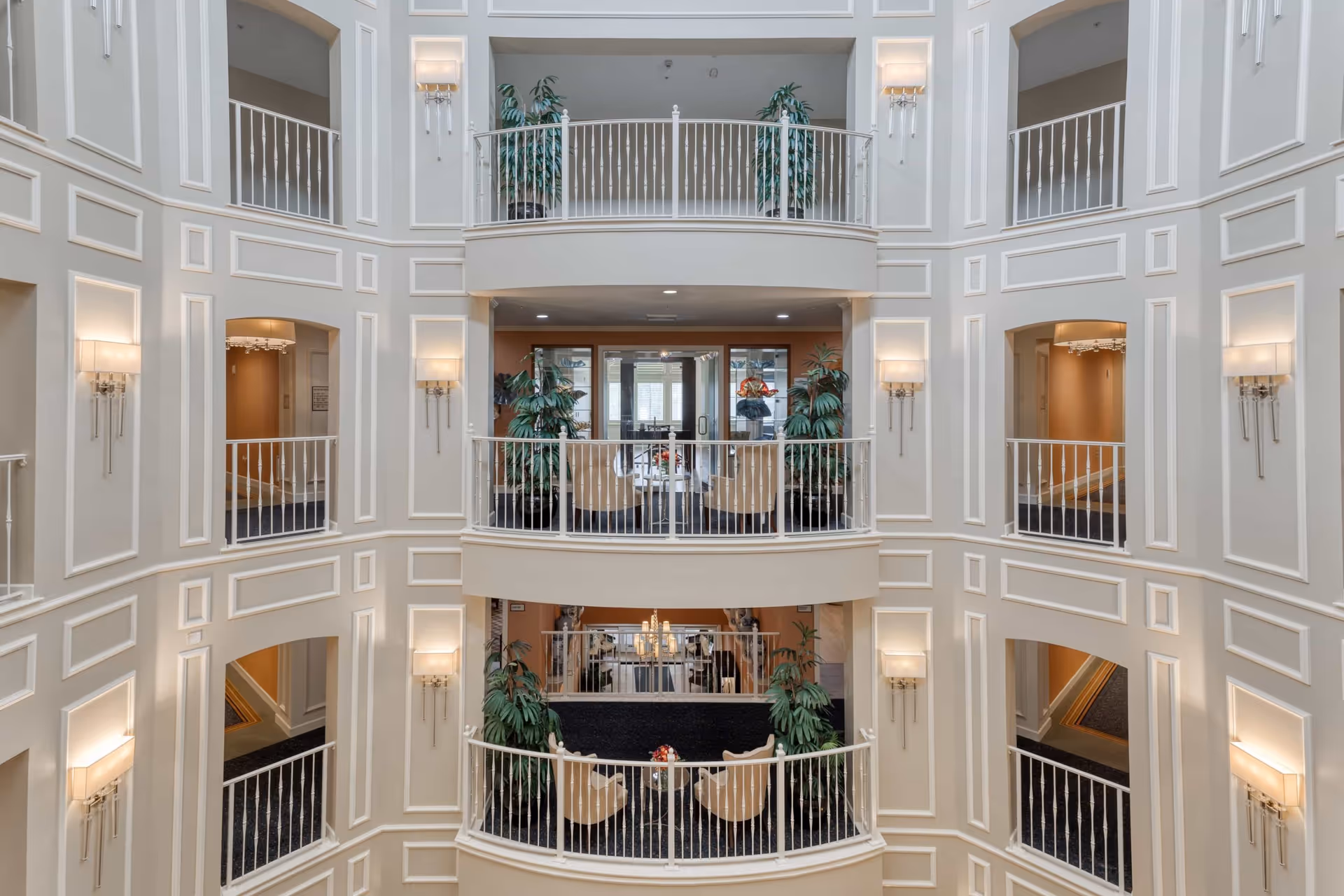 Interior view of a multi-story atrium in a senior living facility with white railings, beige walls with decorative molding, wall sconces, and potted plants. Seating areas with chairs and tables are visible on the balconies overlooking the atrium.