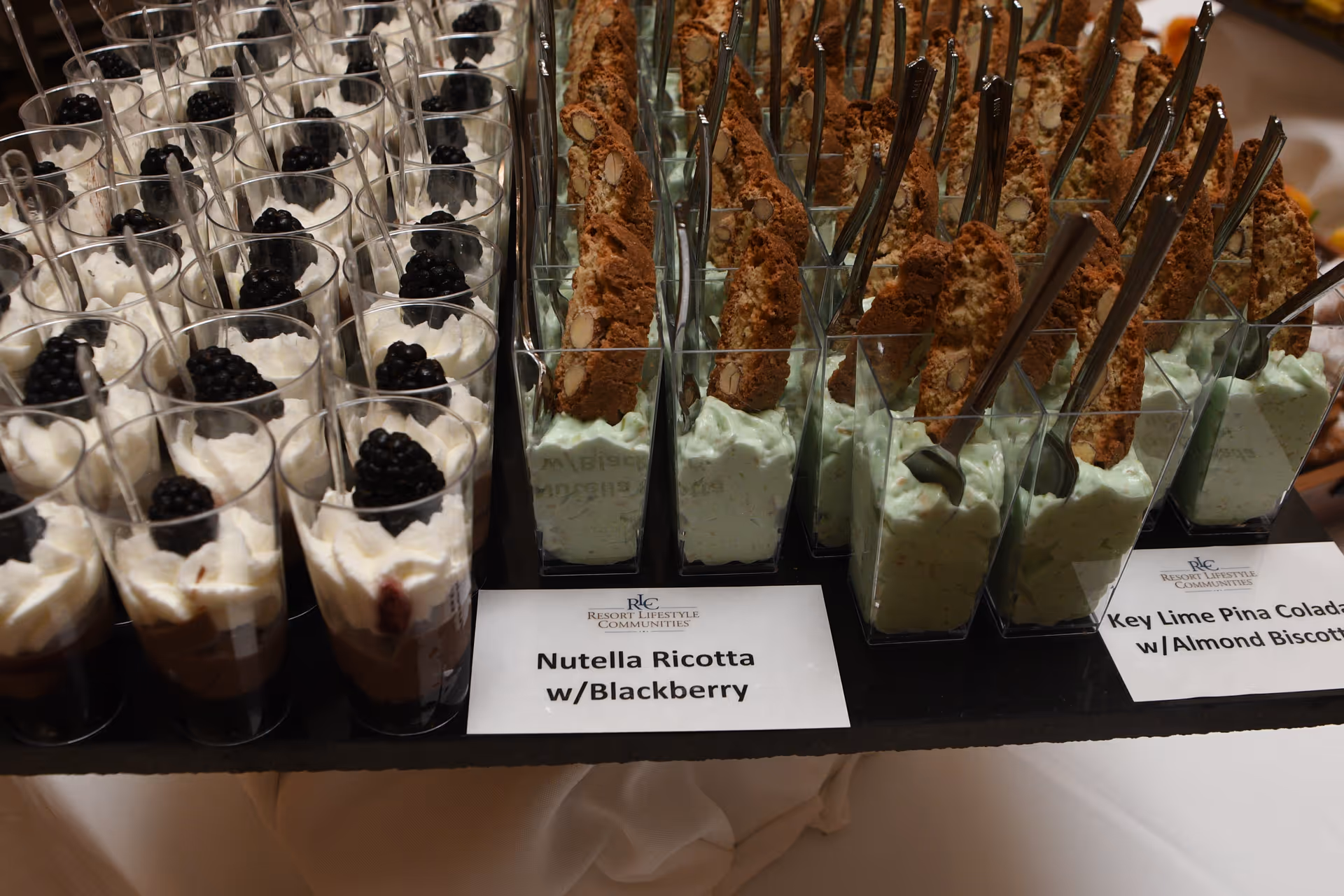 Close-up view of two types of dessert servings in clear plastic cups. On the left, Nutella Ricotta topped with blackberries and whipped cream. On the right, Key Lime Pina Colada mousse with almond biscotti sticks and small spoons. Both desserts are neatly arranged on a black tray with labels in front of them.