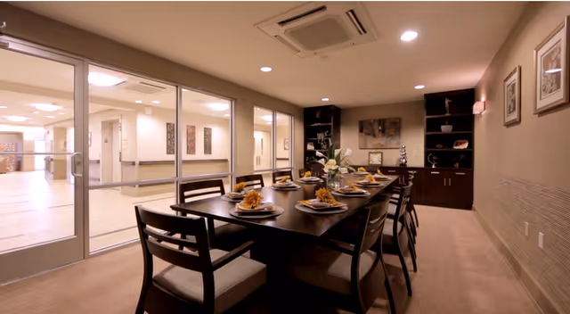 A long dining table set with place settings and chairs in a well-lit interior room with glass doors opening to a corridor.
