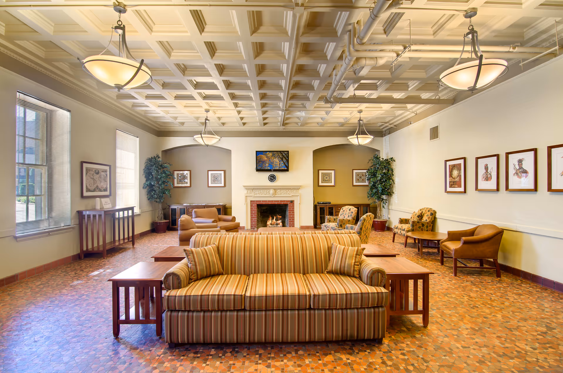 A spacious senior living facility lounge with a striped sofa in the foreground, wooden side tables, armchairs, and a fireplace with a TV mounted above it. The room features a coffered ceiling with hanging light fixtures, large windows with blinds, framed artwork on the walls, and potted plants in the corners.