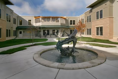 Outdoor courtyard area at Oak Park Place Madison featuring a circular water fountain with a sculpture in the center, surrounded by paved walkways, grass, small trees, and a two-story building with large windows and a pergola on the upper level.