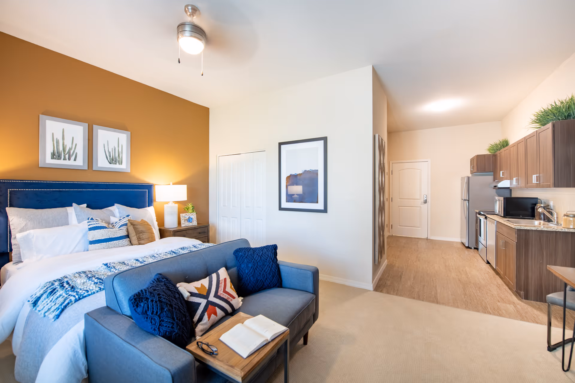 Bright studio-style apartment showing a bed with a navy headboard, a small sofa at its foot, and a kitchenette along the right wall.