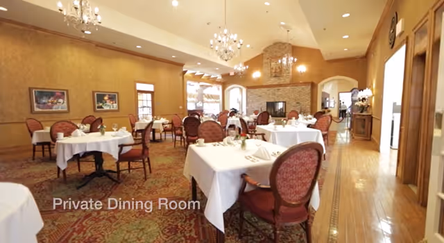 A spacious private dining room with round tables set with white linens, upholstered chairs, chandeliers, and a fireplace.