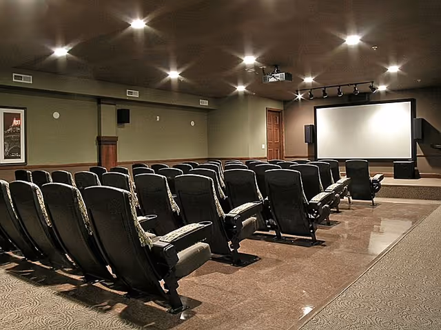 Interior view of a small theater or media room with multiple rows of cushioned seats facing a large projection screen. The room has dim lighting with ceiling spotlights and a projector mounted on the ceiling. The walls are painted in neutral tones and there is a wooden door to the right of the screen.