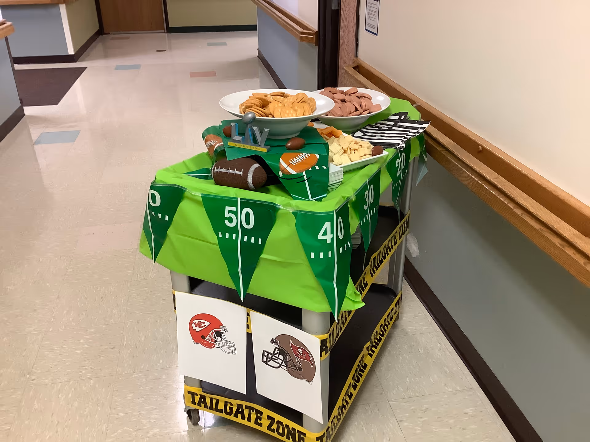 A football-themed snack cart with bowls of crackers, sliced meat, and cheese set in a nursing home hallway.