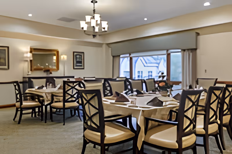 Bright dining room with multiple set tables and cushioned chairs under a chandelier near large windows.