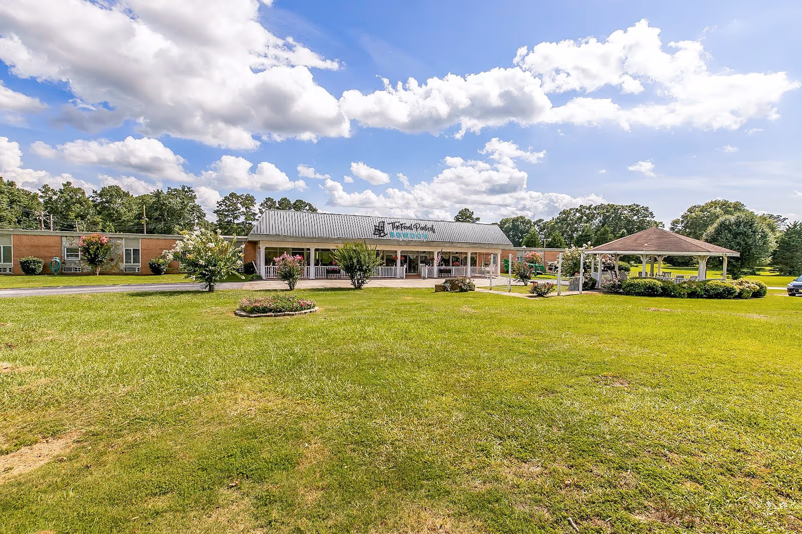 Front exterior view of Front Porch of Bowdon Senior Living facility with a large green lawn, a covered gazebo area to the right, and a partly cloudy blue sky above.