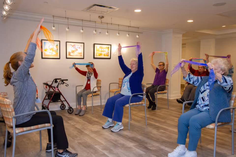 A group of elderly individuals seated in chairs in a well-lit room participating in a seated exercise class using colorful scarves. The instructor, also seated, leads the group with arms raised holding an orange scarf. The room has wooden flooring, framed artwork on the wall, and a walker is visible near the back wall.