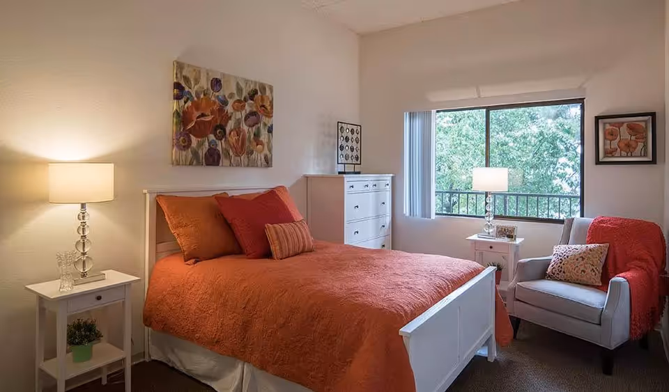 A cozy bedroom with a white bed frame and an orange bedspread, complemented by matching pillows. There are two white nightstands on either side of the bed, each with a lamp. A white dresser is positioned against the far wall. A comfortable armchair with a red throw blanket and a decorative pillow sits near a large window with vertical blinds, allowing natural light to brighten the room. The walls are adorned with floral artwork.