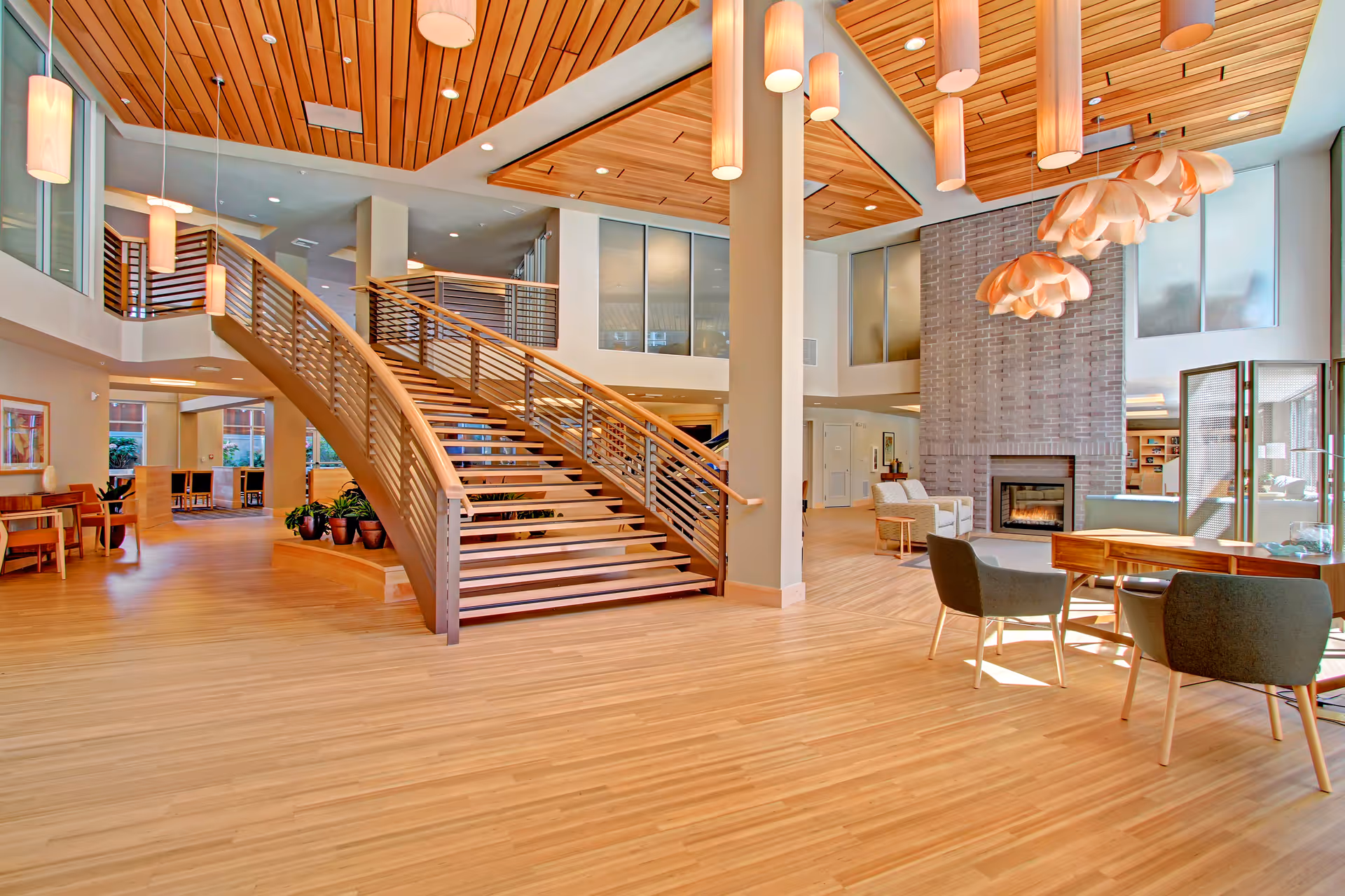 Bright open two-story lounge with a central wooden staircase, seating areas, fireplace, and modern pendant lights.
