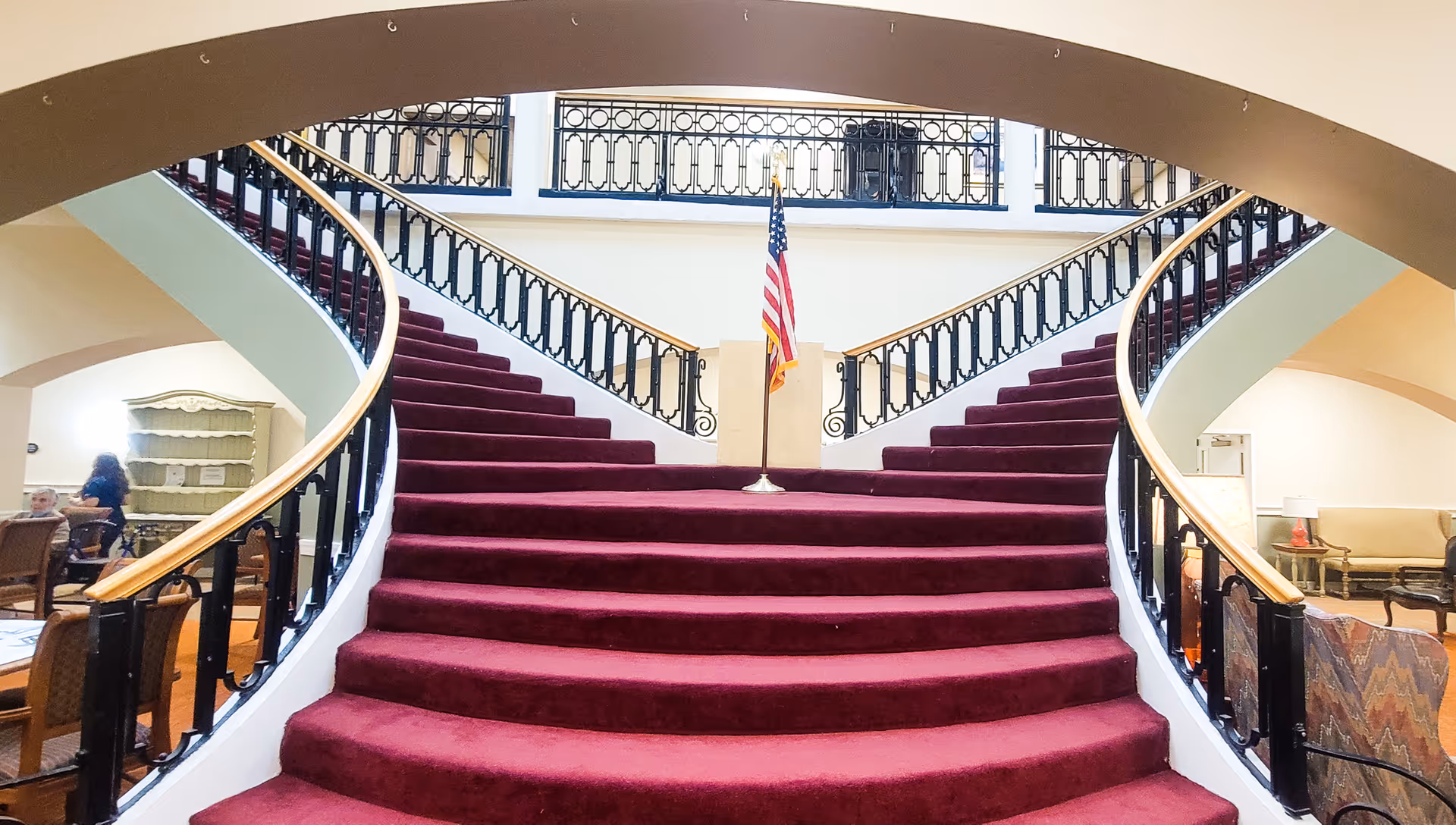 Wide, curved staircase with red carpet leading up to a landing with an American flag in the center. The staircase has black railings with wooden handrails. On either side of the staircase, there are seating areas with chairs and tables. The space is well-lit with natural light coming from above.