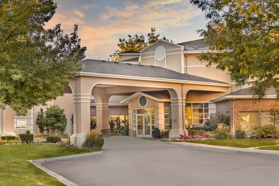 Exterior view of Legacy Retirement Residence building entrance with a covered driveway, surrounded by trees, shrubs, and flowers under a partly cloudy sky at sunset.