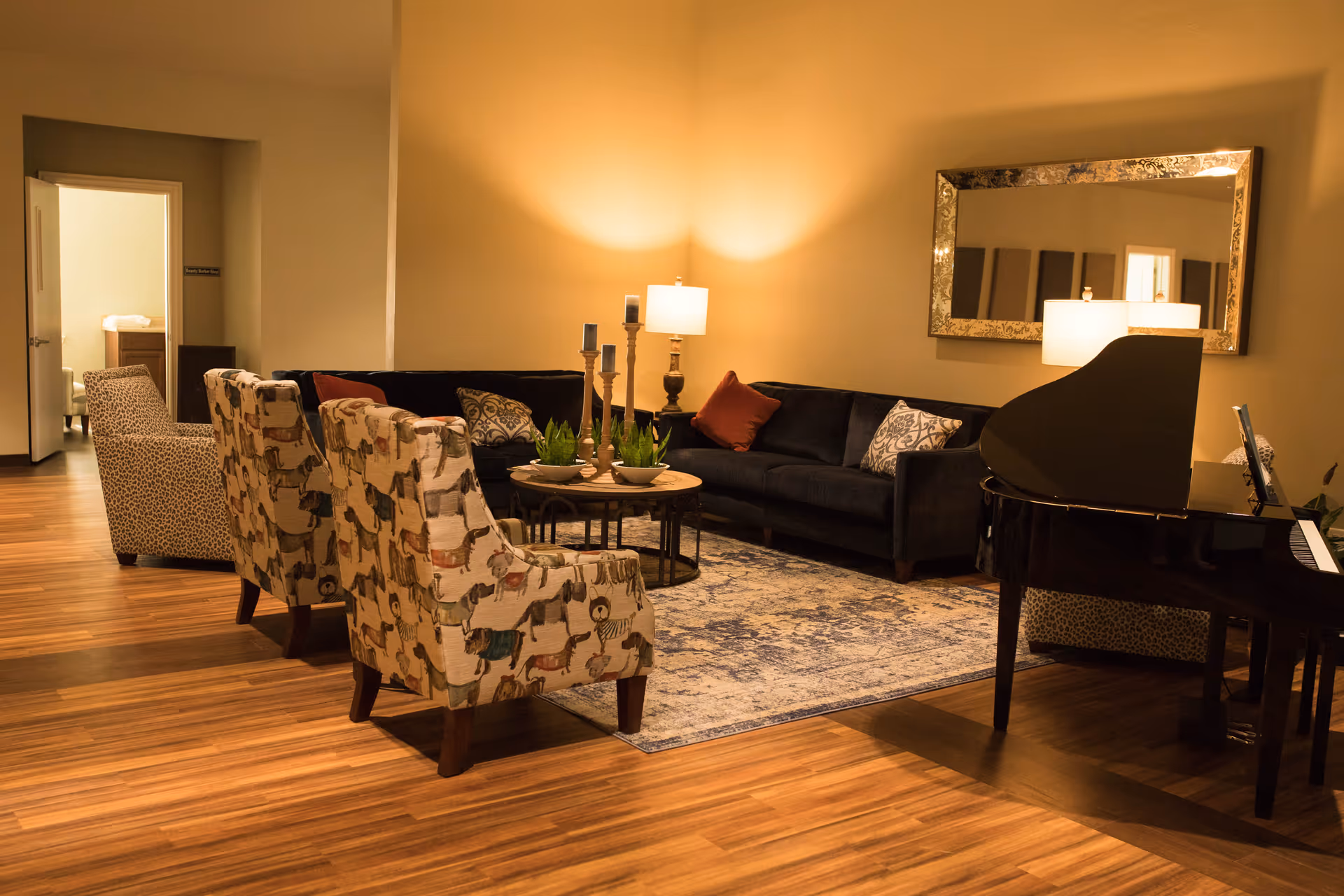 A cozy living room area with wooden flooring, featuring a black grand piano on the right, a large dark sofa with patterned and solid pillows, two patterned armchairs with animal prints, a round coffee table with plants and candles, a large decorative mirror on the wall, and warm lighting from table lamps.