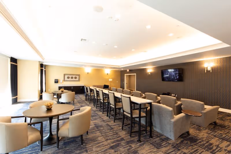A spacious senior living common area with multiple seating options including round tables with beige chairs, a long table with high chairs, and a row of armchairs facing a wall-mounted TV. The room has a patterned carpet, soft lighting from wall sconces, and large windows with curtains allowing natural light.