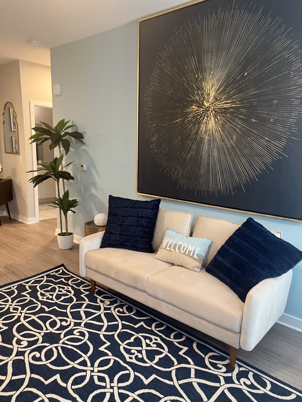 A cozy living room area with a beige sofa adorned with two large dark blue pillows and a smaller pillow with the word 'WELCOME'. Behind the sofa is a large piece of wall art featuring a gold starburst design on a dark background. A patterned navy and white rug covers the floor, and a tall green plant in a white pot is placed near the wall. The room has light-colored walls and wood flooring.