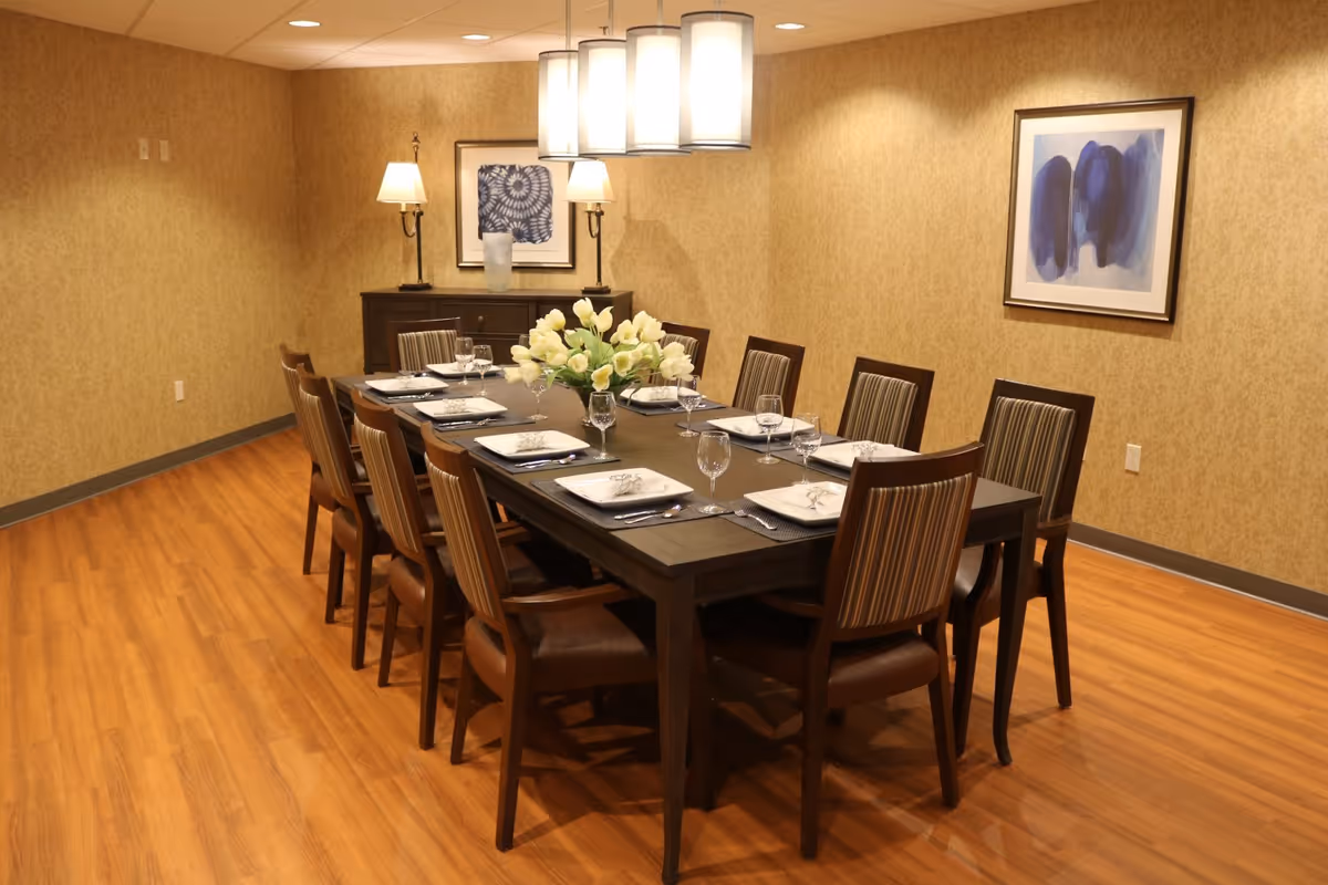 A dining room with a long dark wooden table set for ten people. Each place setting includes a white square plate, a napkin with a napkin ring, a fork, a spoon, and a wine glass. A centerpiece of white flowers is in the middle of the table. The room has wooden flooring, beige patterned wallpaper, two framed abstract paintings on the walls, and two table lamps on a sideboard against the back wall. A modern ceiling light fixture with four cylindrical lamps hangs above the table.