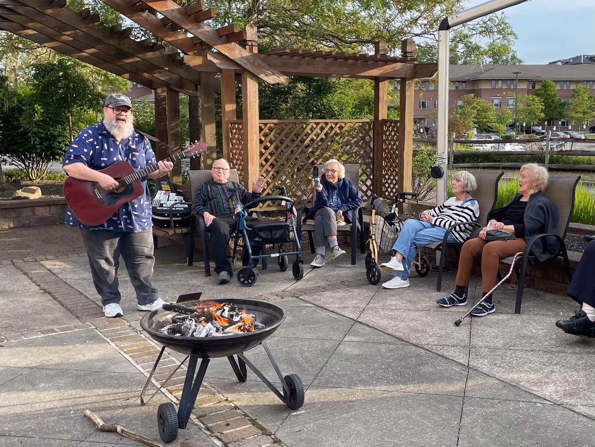 An outdoor patio area at The Villas at King David with a man playing a guitar near a fire pit. Four elderly women are seated on chairs under a wooden pergola, enjoying the music and the fire. The background shows greenery and a building.